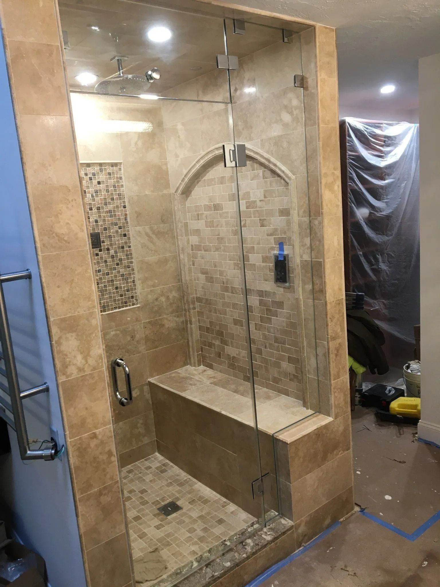 Glass-enclosed shower with beige tile, bench, and arched alcove. Chrome hardware, open door.