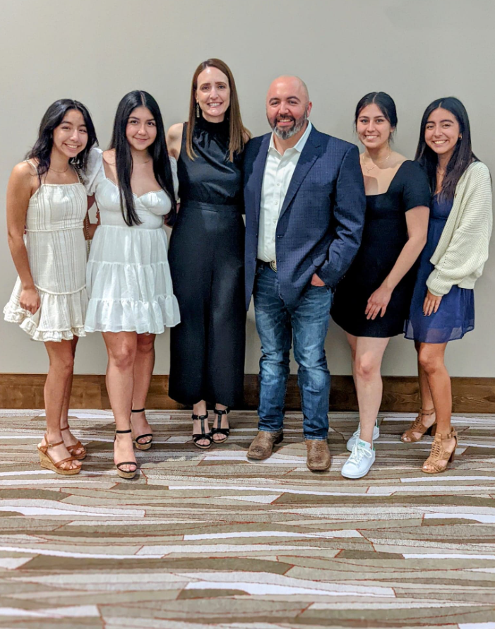 Family group stands together for a photo; two women in white dresses, four in black, patterned wood floor.