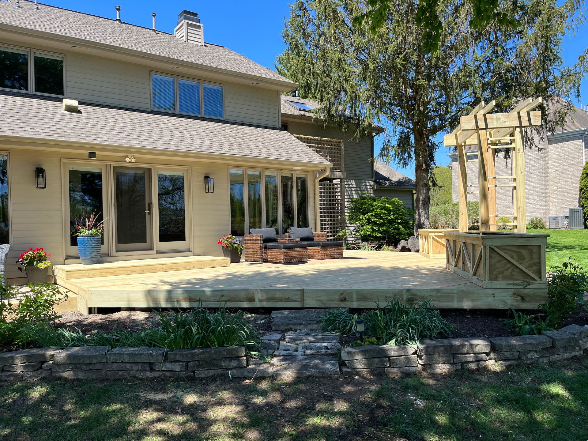 Backyard deck with wooden planters and pergola, adjacent to a beige house, sunny day.