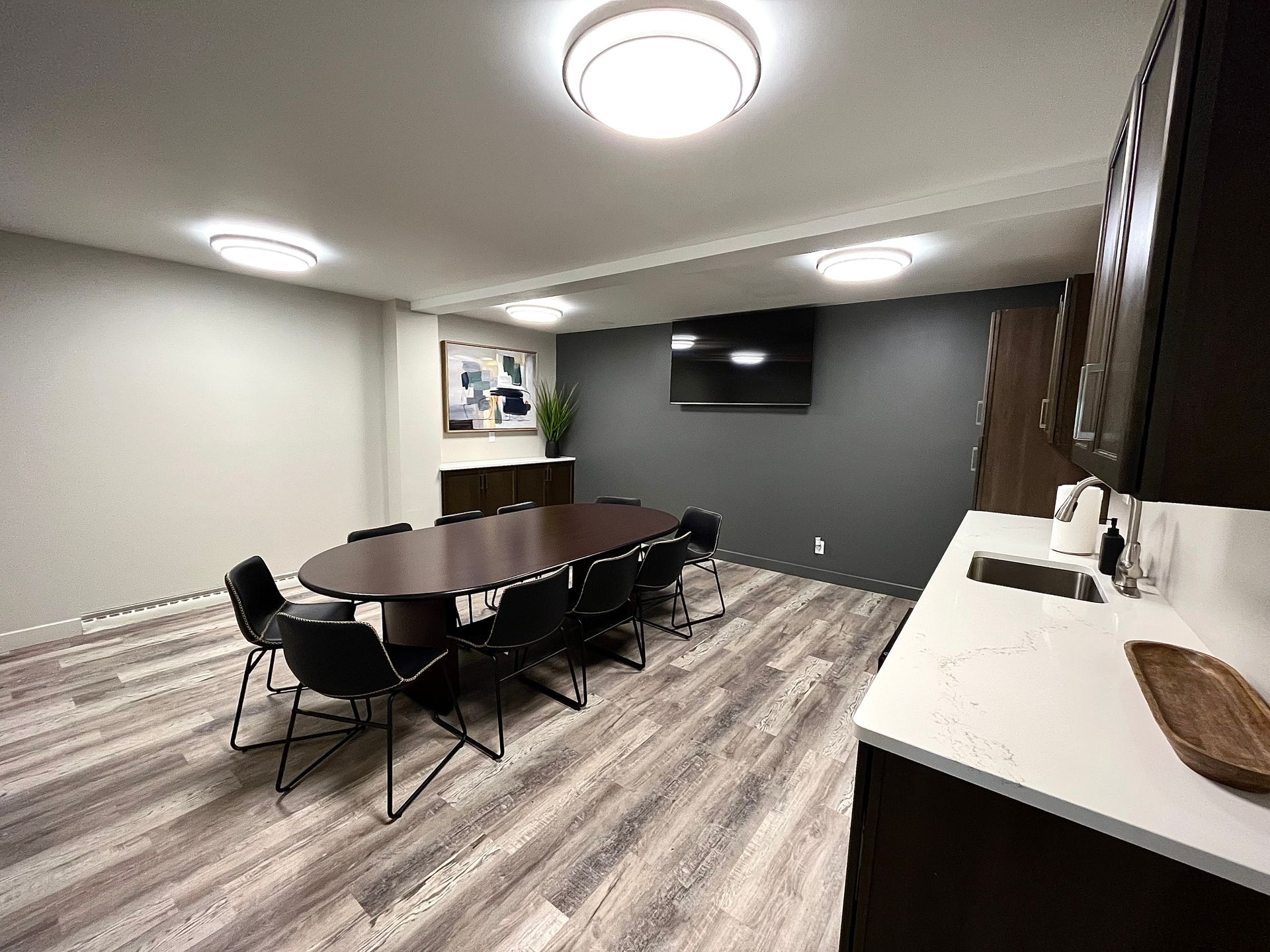 A conference room with a large oval table, chairs, TV, and kitchenette. Modern design with neutral colors.