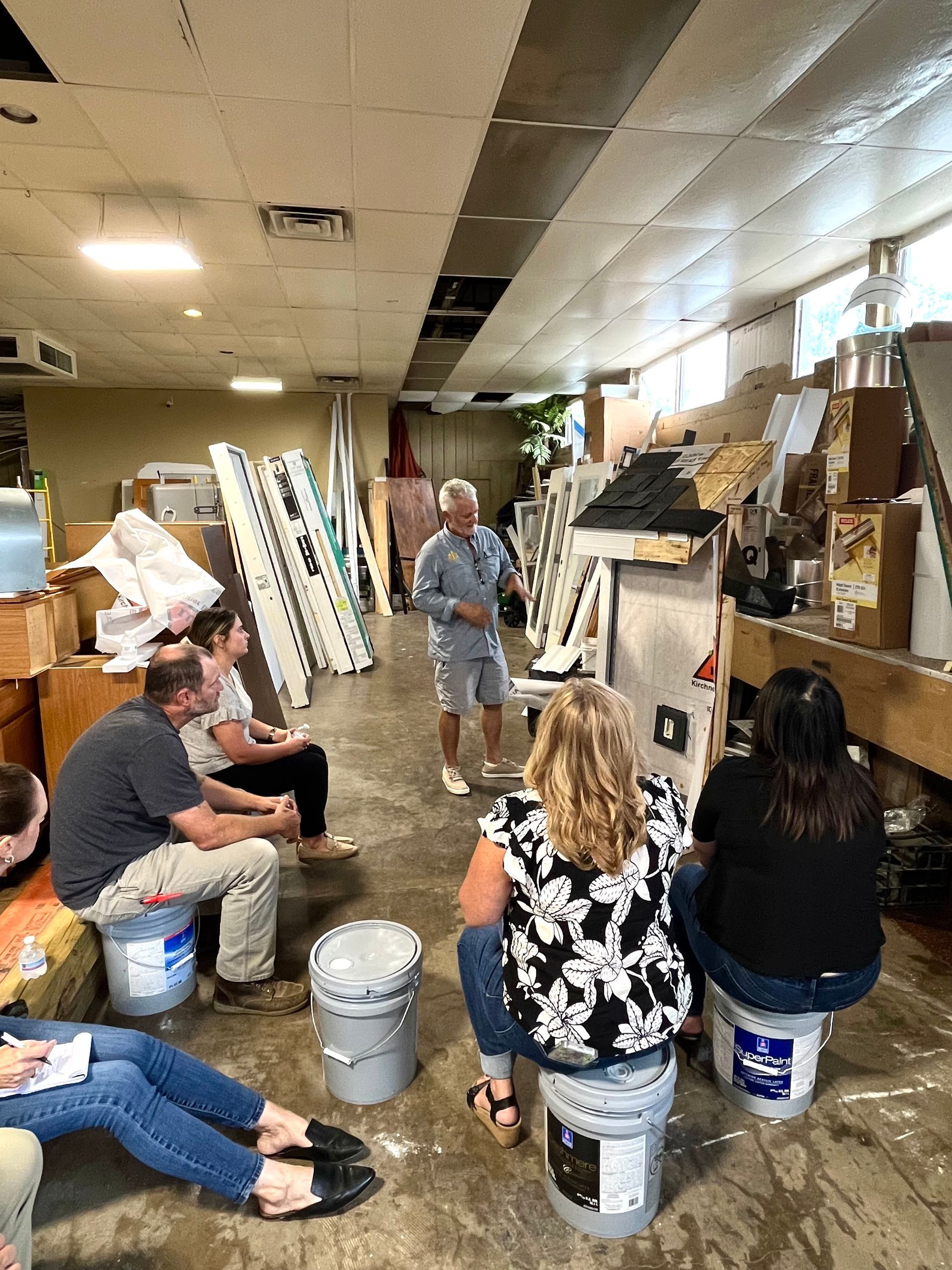 A group seated on buckets listens to a person speaking in a cluttered indoor space.