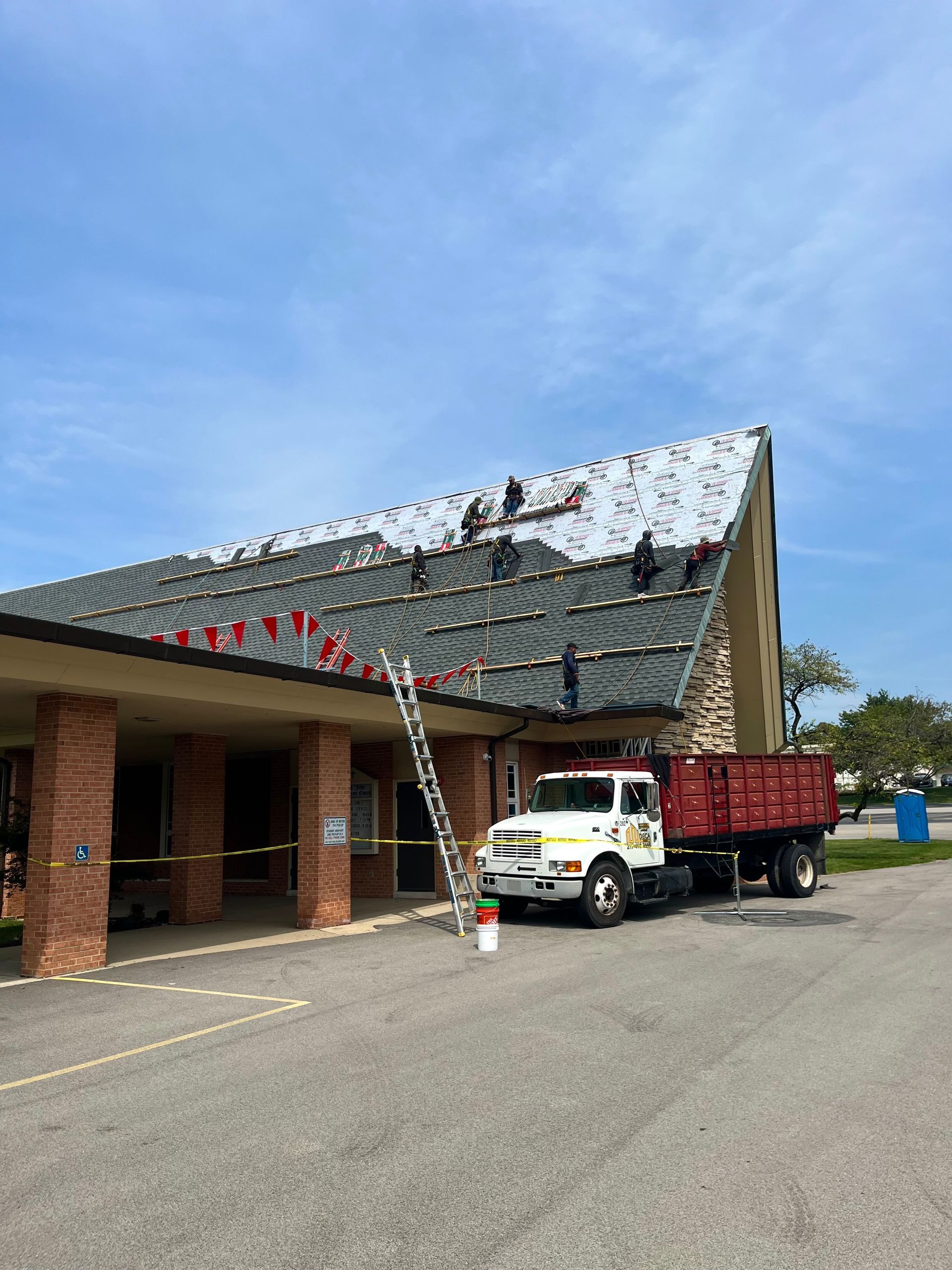 Workers on a roof, replacing shingles. A truck is parked in front of the building.