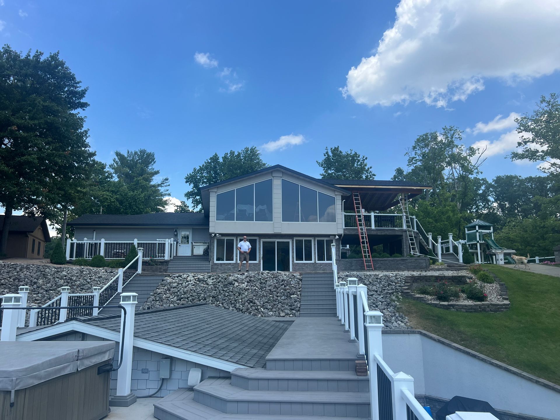 Lakefront house with large windows, stairs, and a raised patio, under a blue sky.