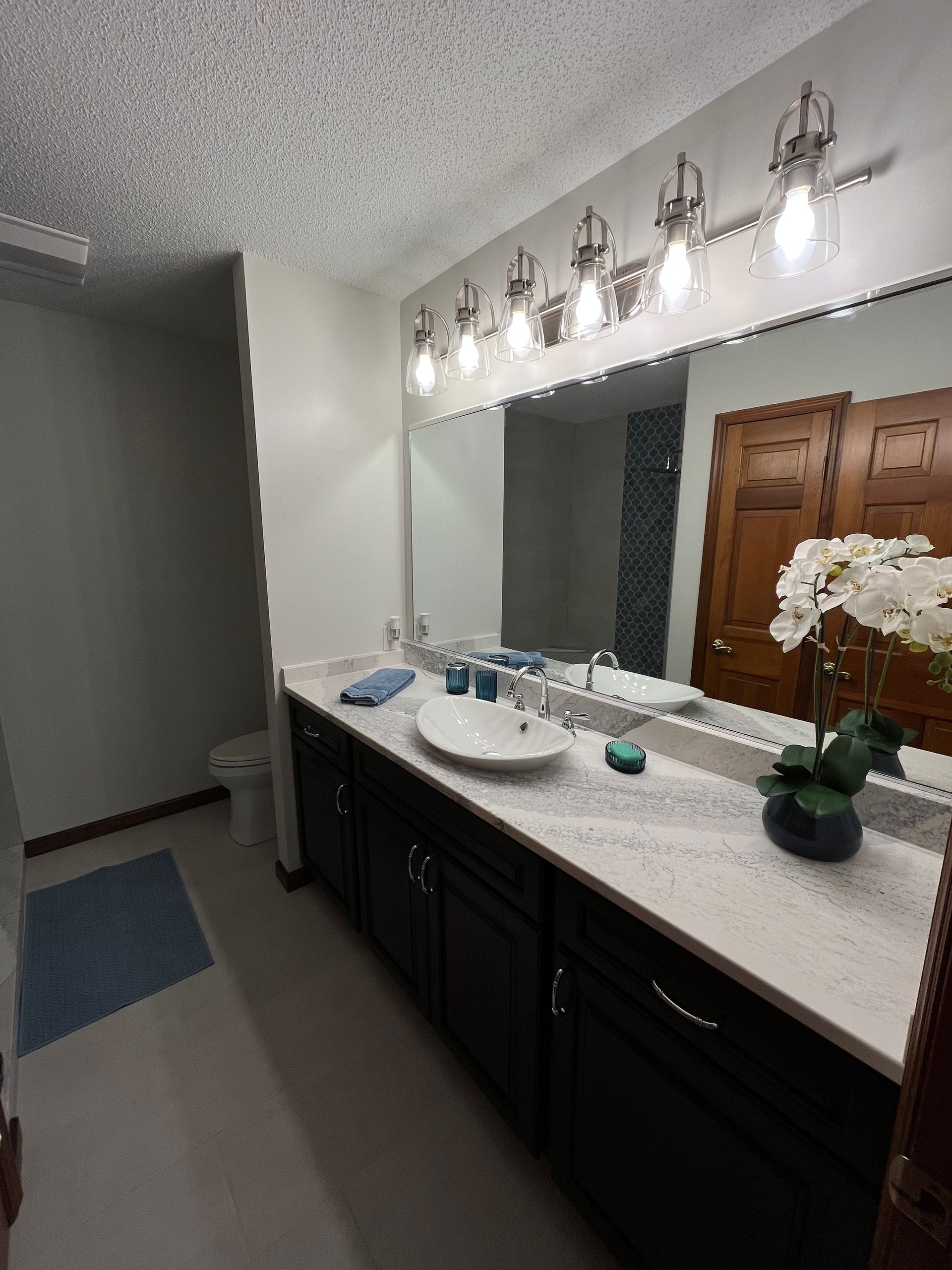 Bathroom with a dark vanity, white countertop, and large mirror. Overhead lights and a decorative flower arrangement.