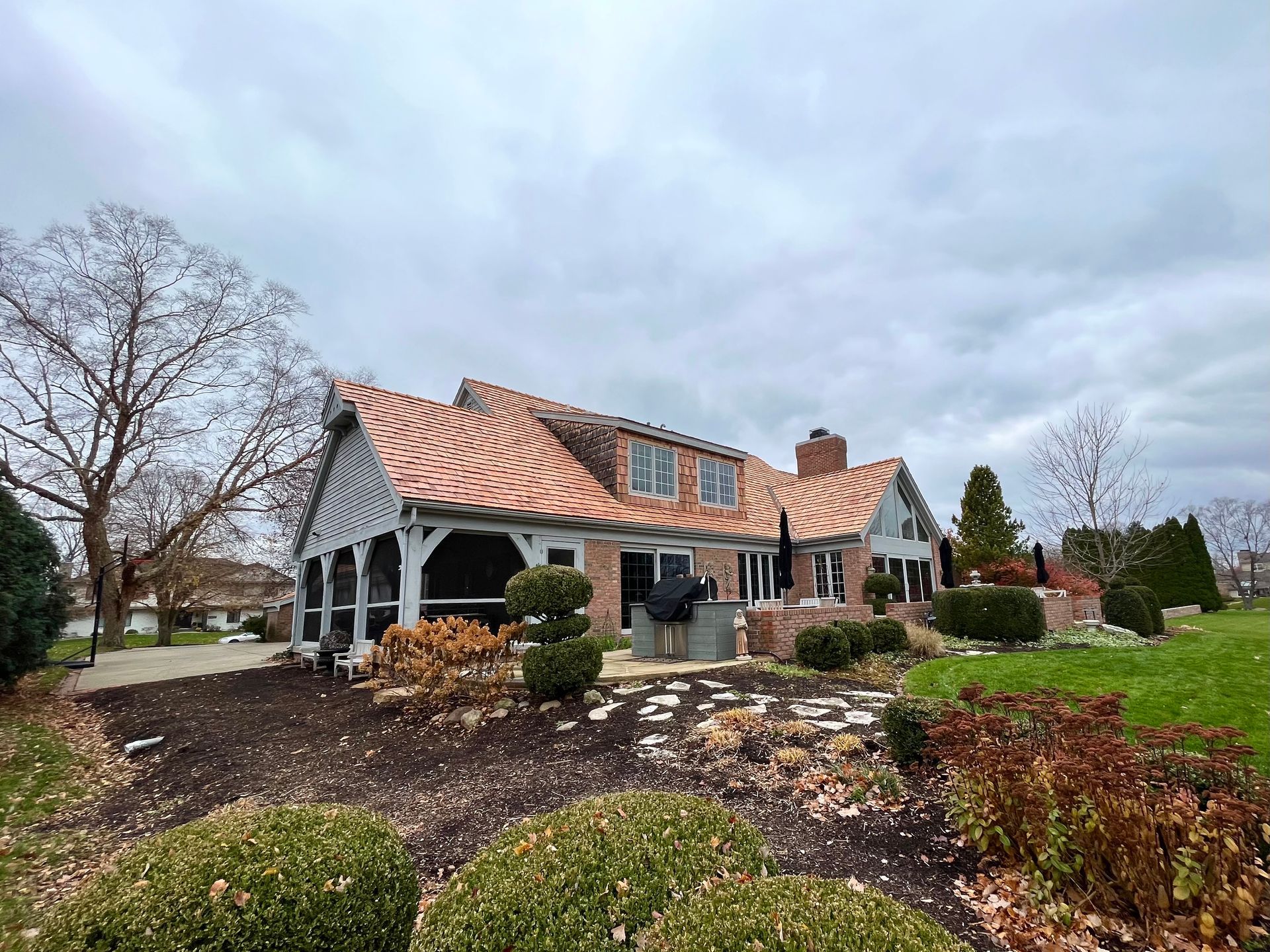 Rear view of a brick house with a copper roof under a cloudy sky, surrounded by landscaping.