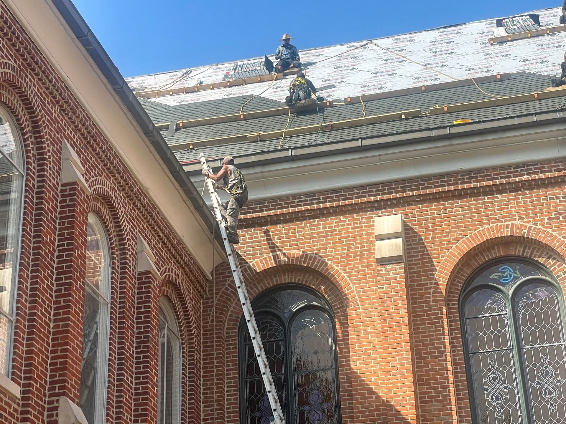 Workers on a church roof repair shingles; a ladder leans against the brick wall.