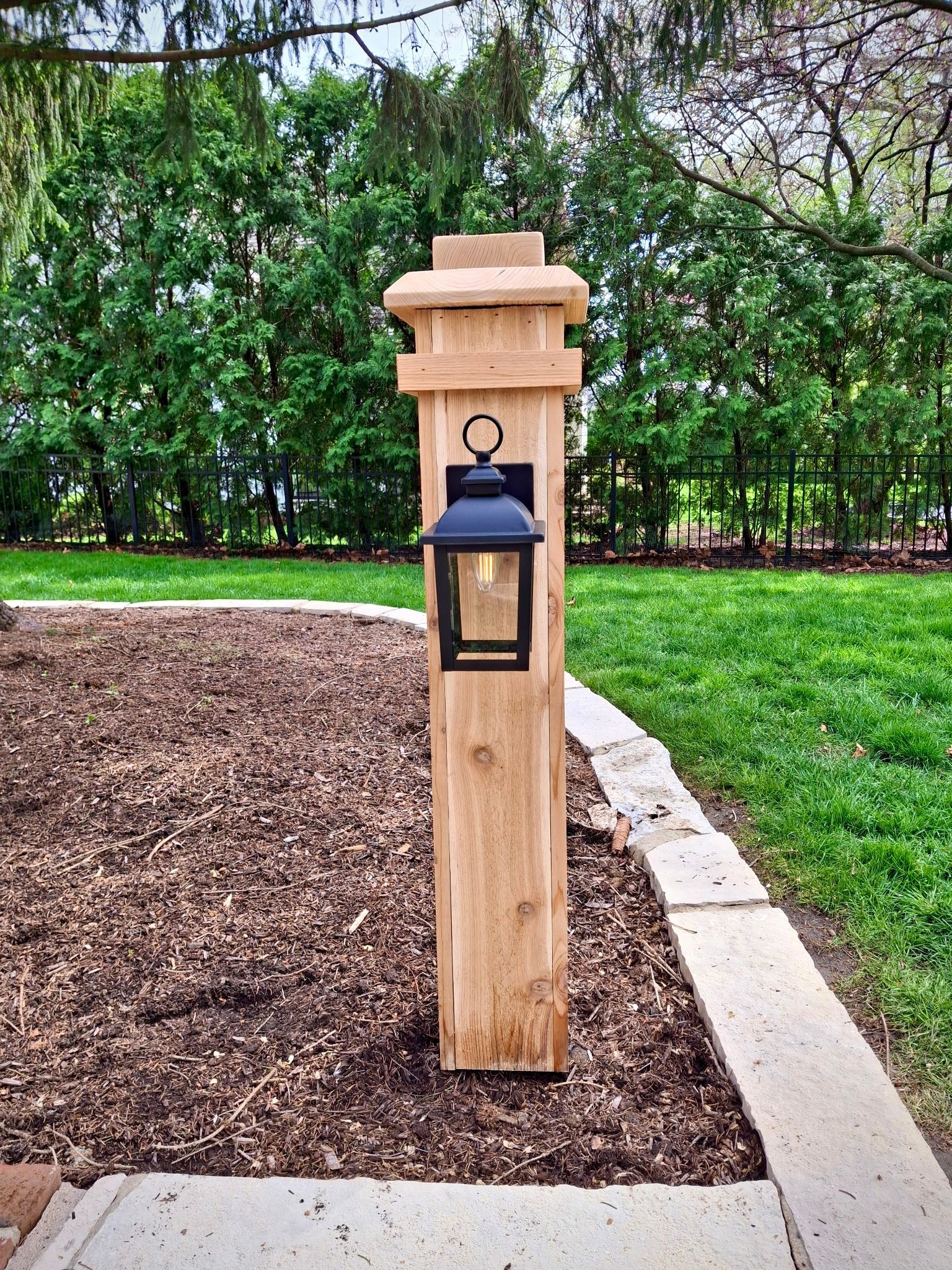 Wooden post with a black lantern, set in a mulch bed, with a grassy lawn and trees in the background.