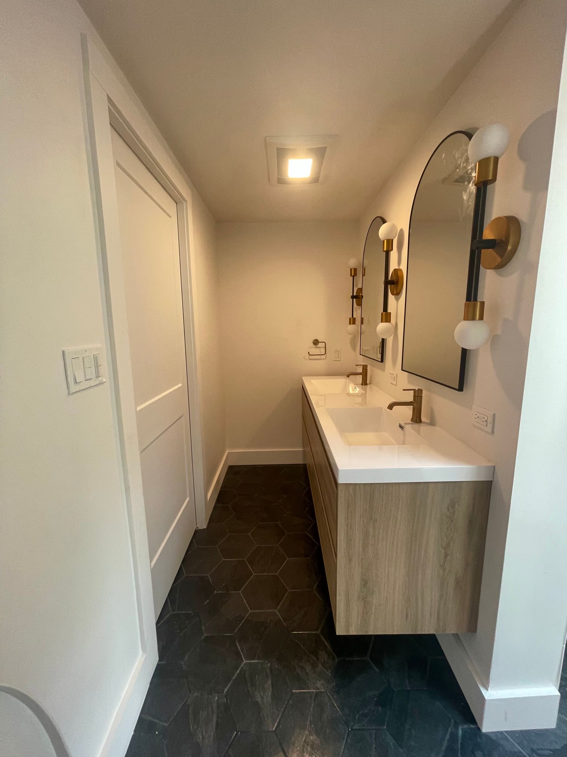 Bathroom with a wooden vanity, two arched mirrors, gold fixtures, and dark tile floor.