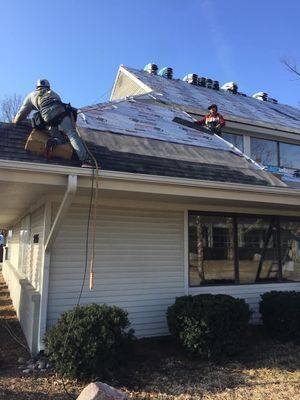 Two workers replacing shingles on a rooftop; daylight, clear sky. One uses a blower.