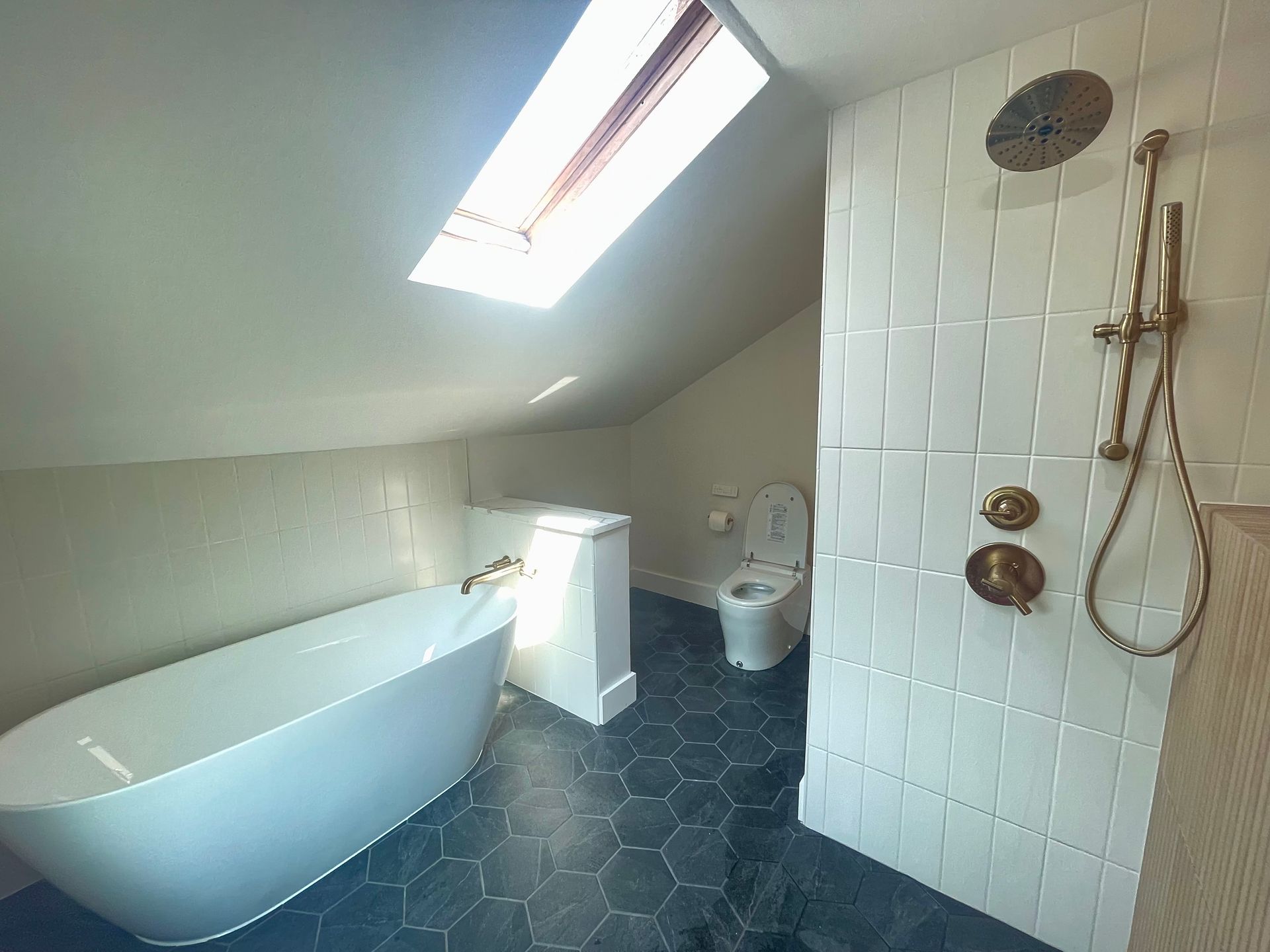 Bathroom with a soaking tub, toilet, and shower with brass fixtures under a skylight.