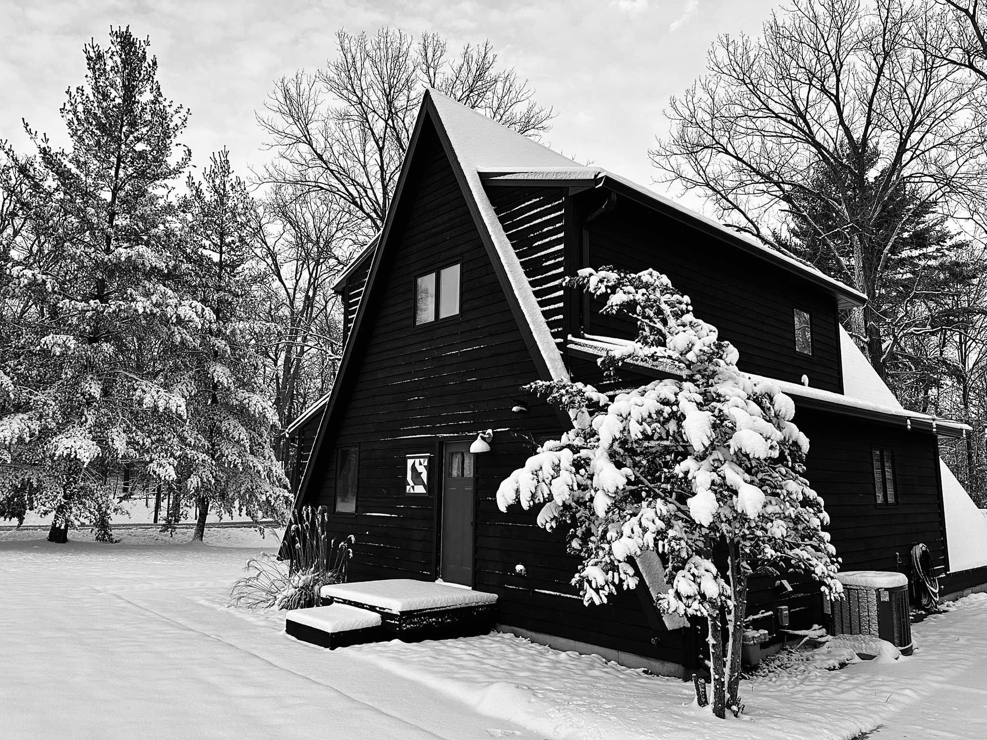 Black and white photo of snow-covered A-frame house in a winter landscape with evergreens and bare trees.