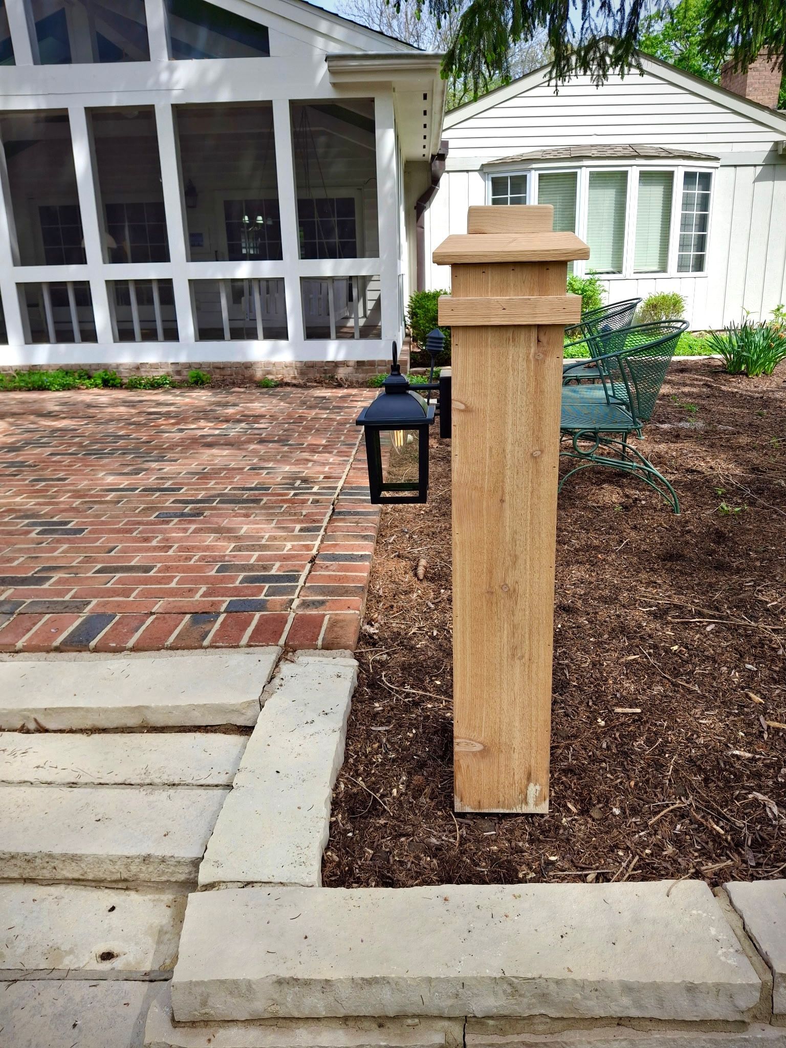 Wooden post with a hanging lantern in front of a brick patio and a white house.