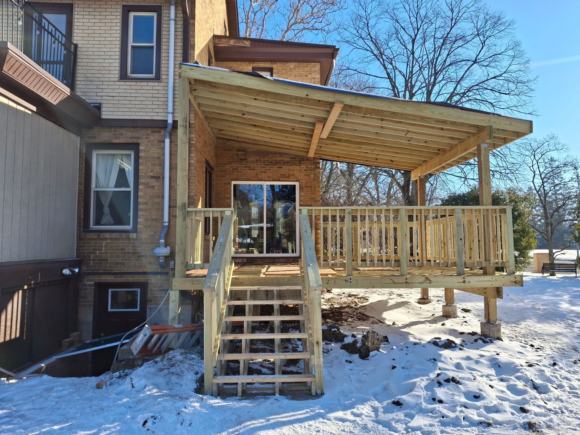 Wooden deck with stairs, attached to a brick building, covered by a sloped roof. Snow covers the ground.