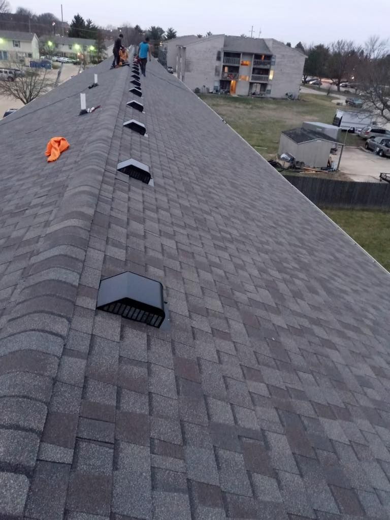 Workers on a gray shingled roof installing black vents, with buildings and a parking lot in the background.