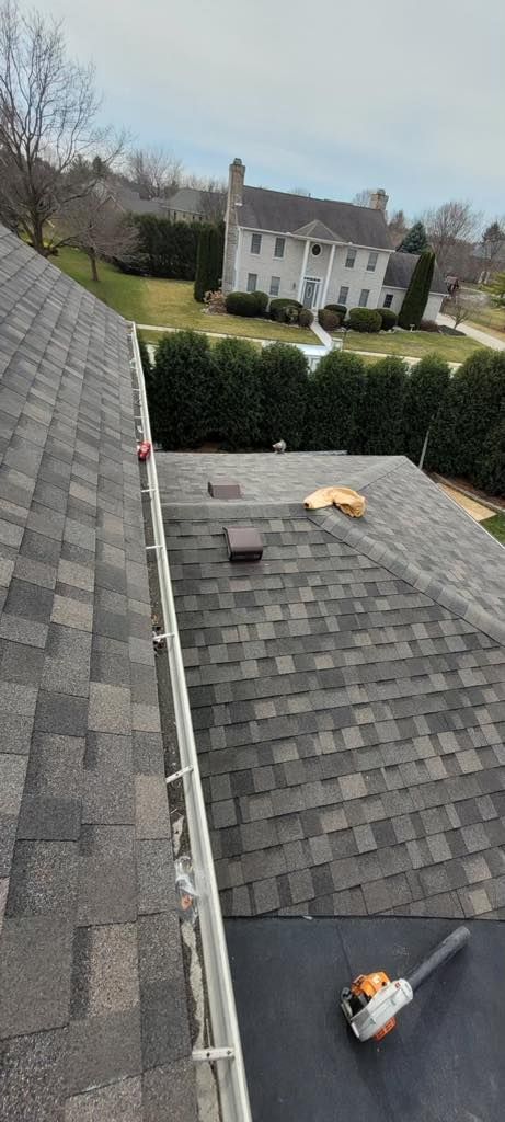 View from a roof, showing shingles, a gutter, and a house in the background. A chainsaw lies on the roof.