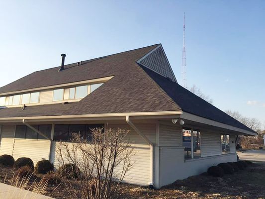 Low-angle view of a light gray building with a dark roof. Bare bushes in front. A radio tower is in the background.