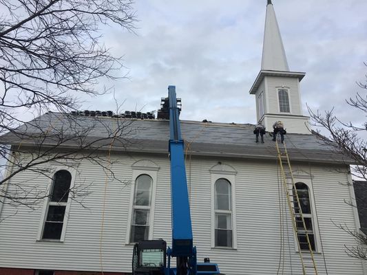 Workers on a church roof with a blue lift and ladder; overcast sky.
