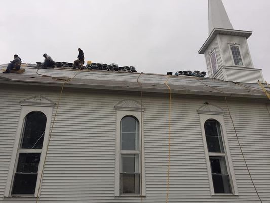 Workers replacing the roof of a white church with arched windows and a steeple. Cloudy sky.