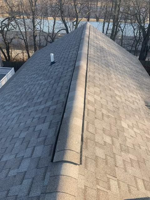 View of a gray asphalt shingle roof with a ridge vent, a white vent pipe, and trees in the background.