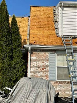 Roof partially stripped of shingles, showing exposed wood, next to a brick building. Ladder leans against the building.