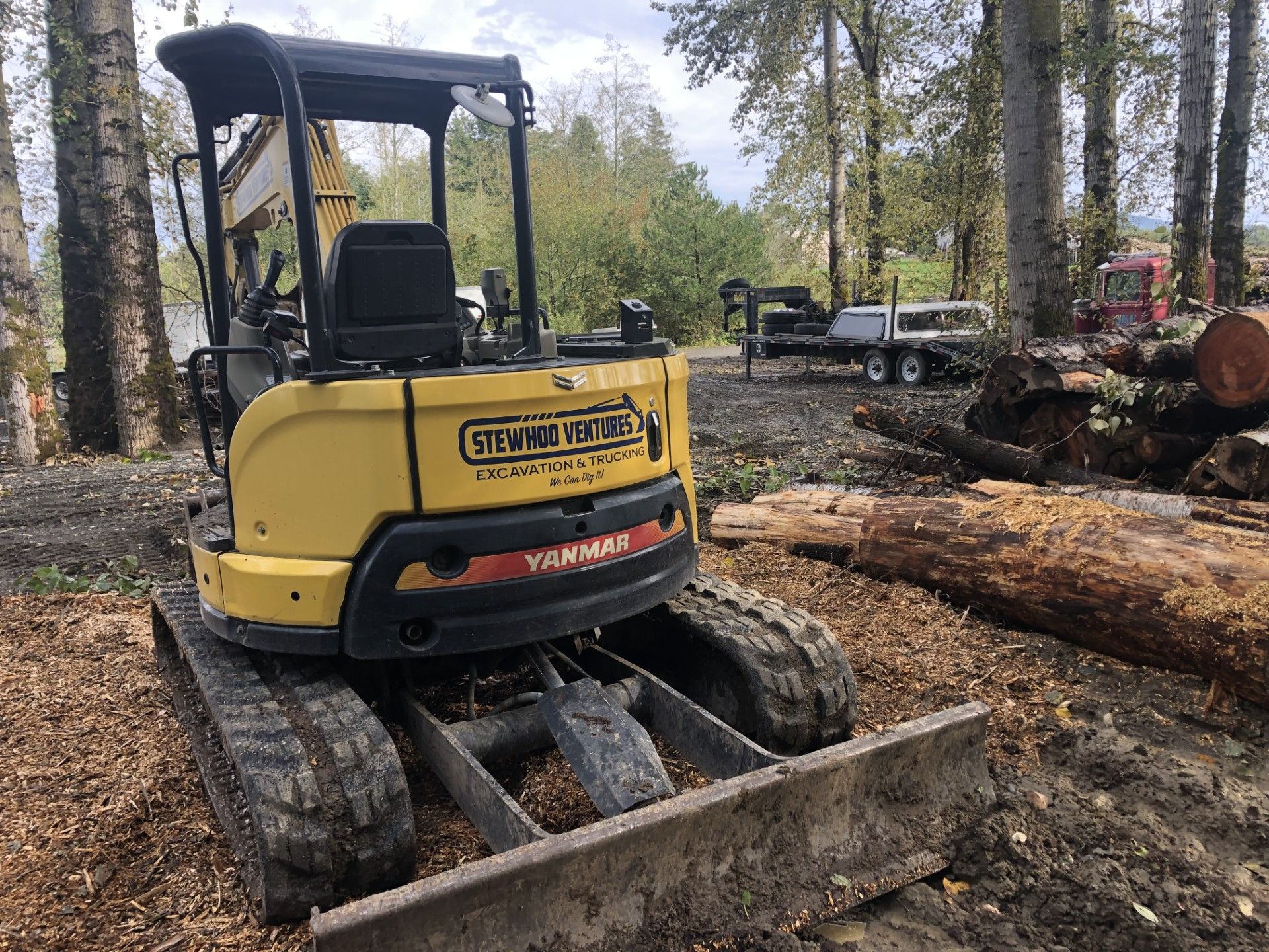 A yellow excavator is parked in a forest next to a pile of logs.