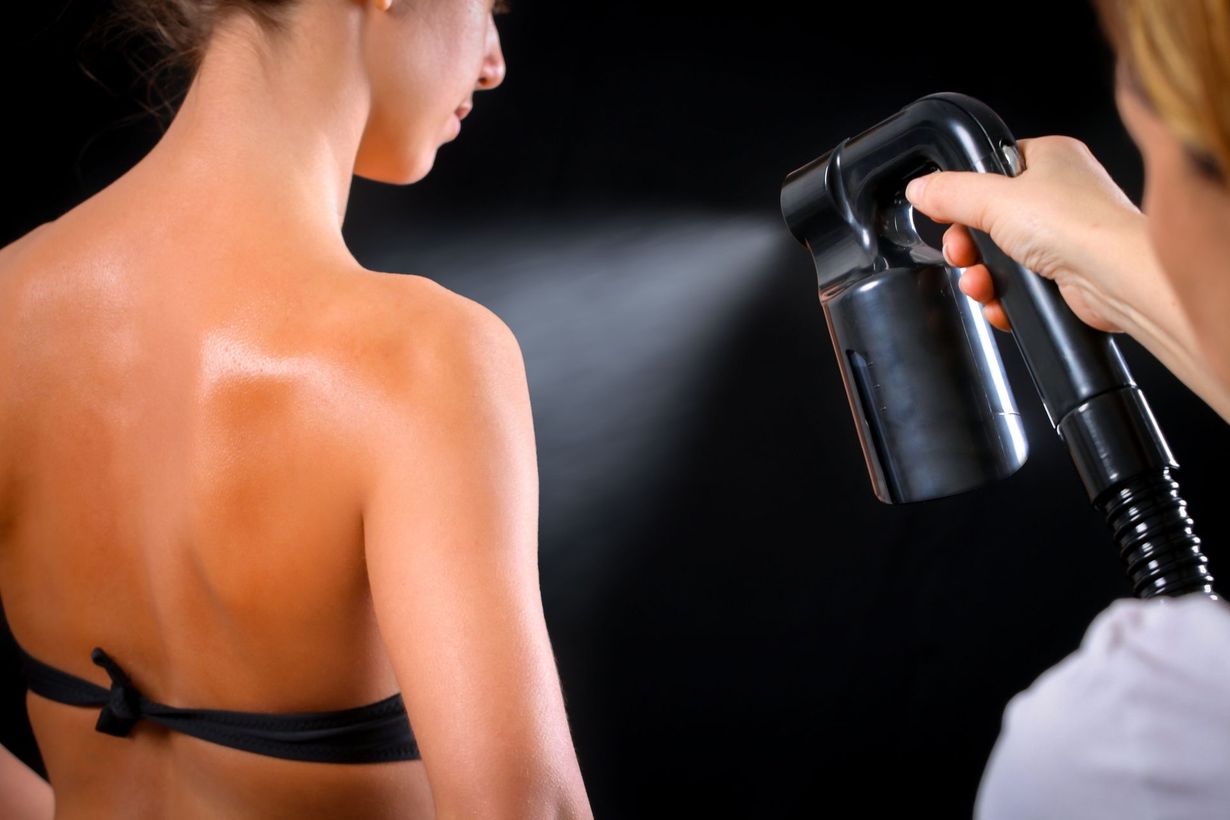 Woman's back being spray tanned by another person with a dark spray gun.