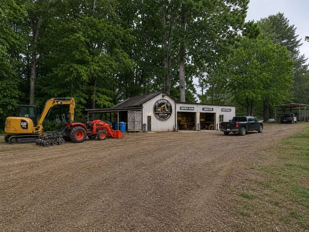 Gravel lot with a yellow excavator, red tractor, white shed, and black pickup under trees