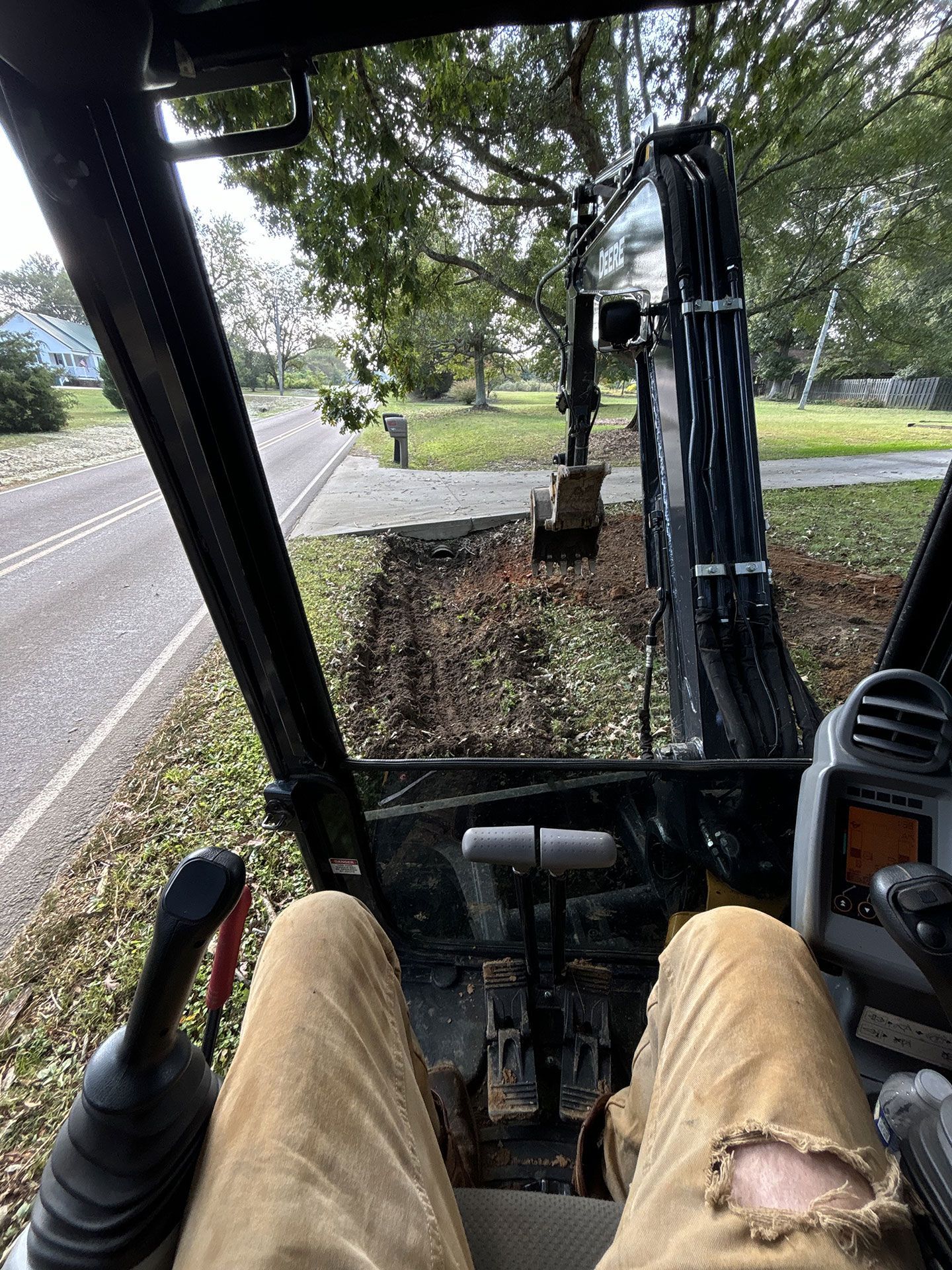 Inside a construction vehicle cab, legs visible, facing a tree-lined road and grassy park area.