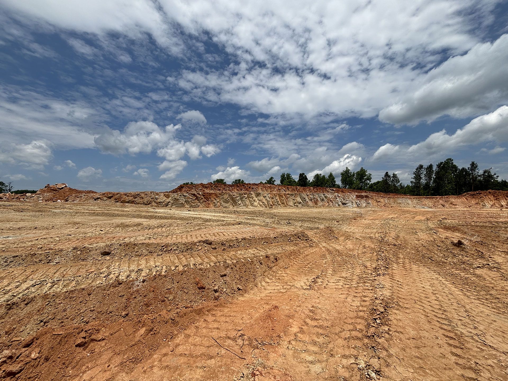 Wide dirt clearing under a cloudy sky with tire tracks and trees on the horizon