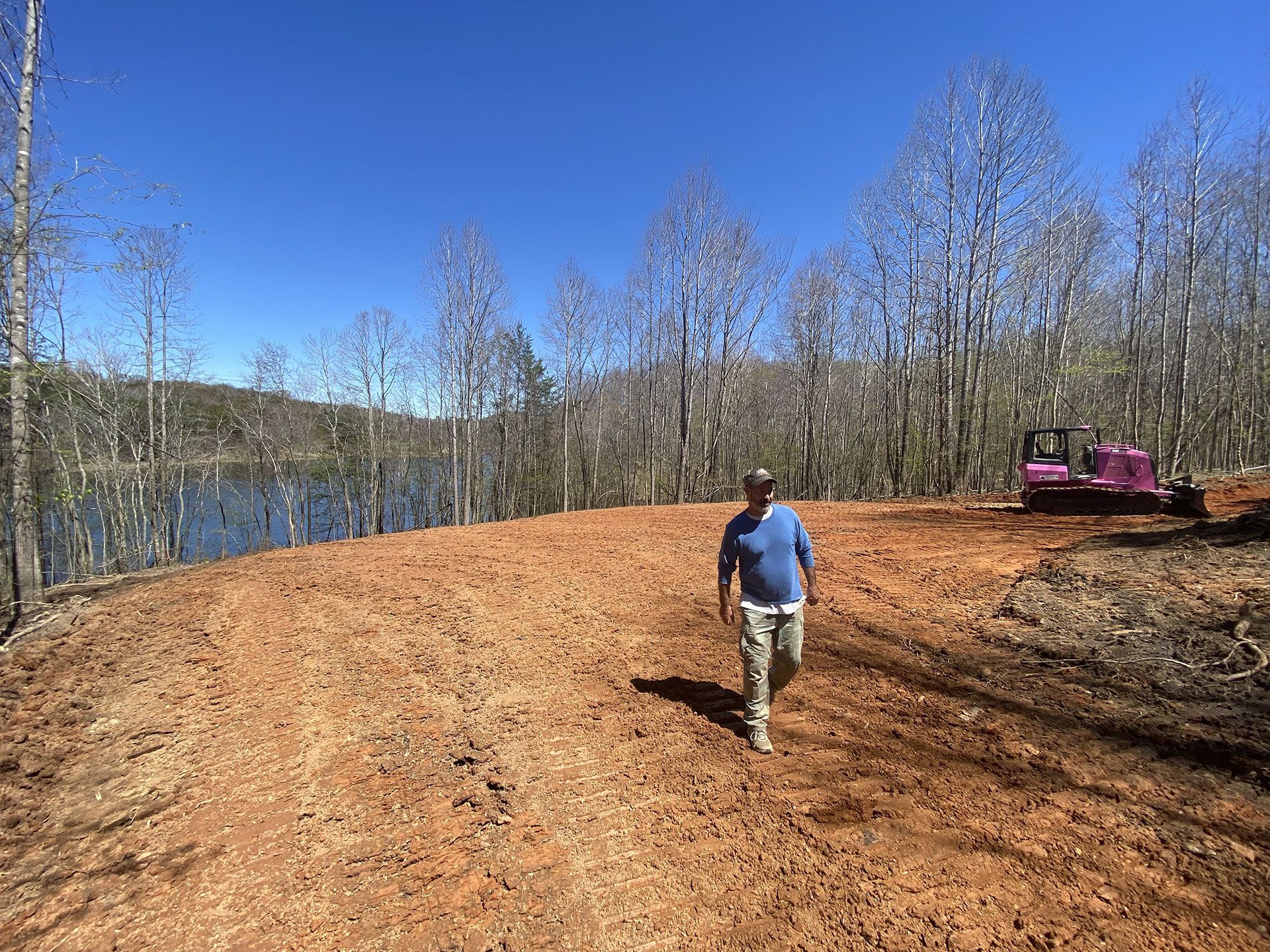 Person walking on a dirt road by a lake, with bare trees and a red tractor nearby.