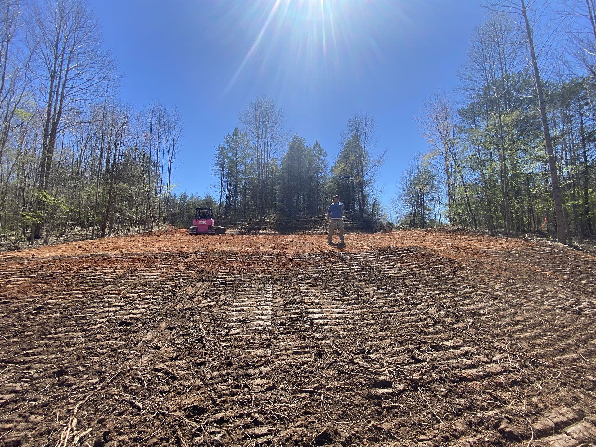 Dirt field under a bright blue sky, with two small people and a pink object near the treeline.