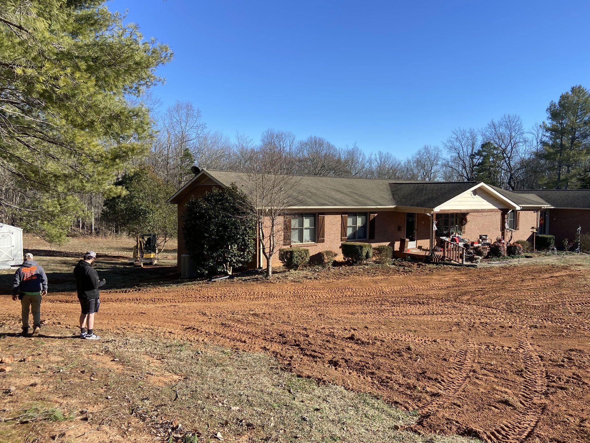 Rural brick house with a muddy yard; two people walk at left under a clear blue sky.