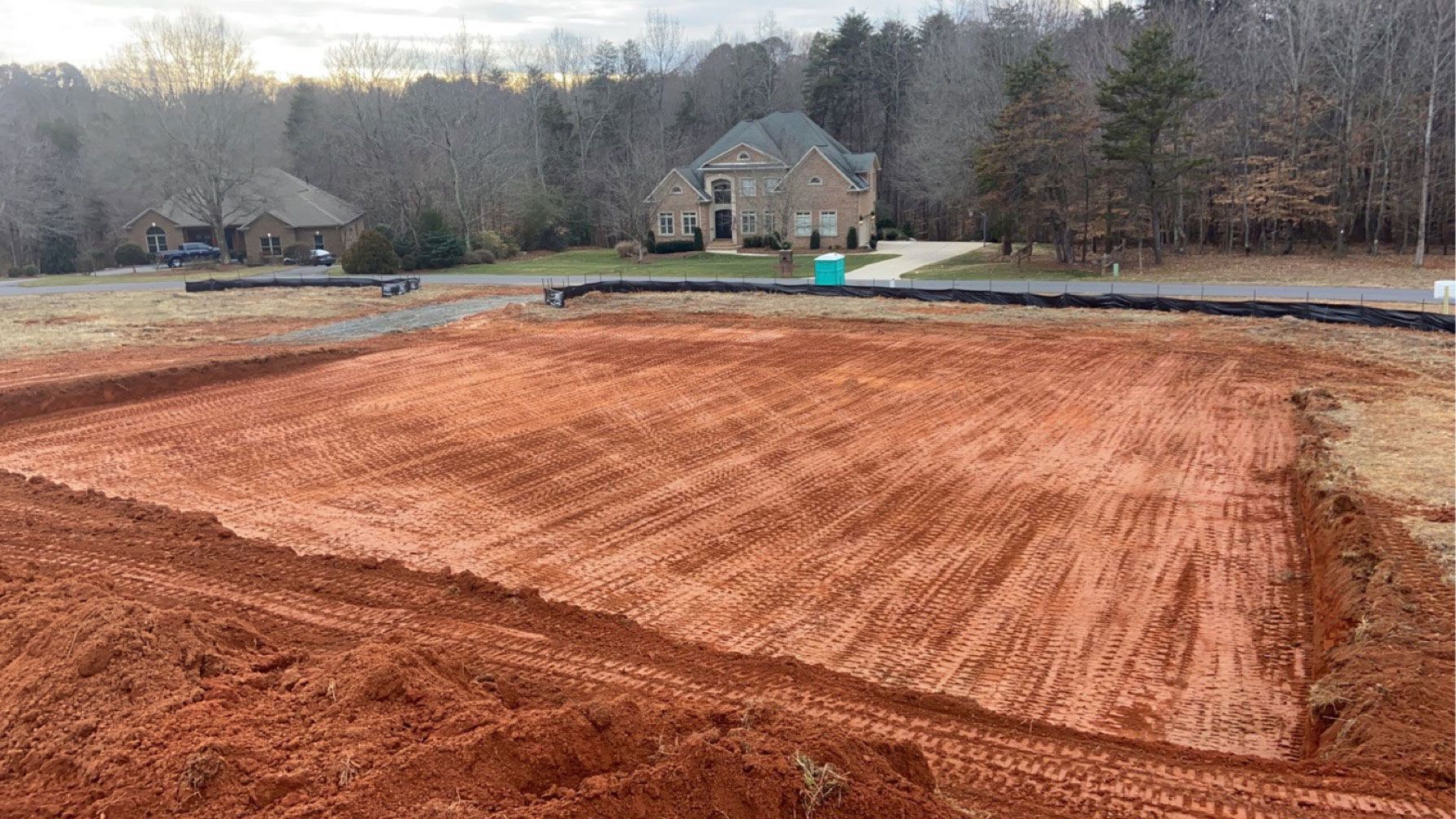 Freshly graded red dirt lot near a house, with trees and a cloudy sky in the background