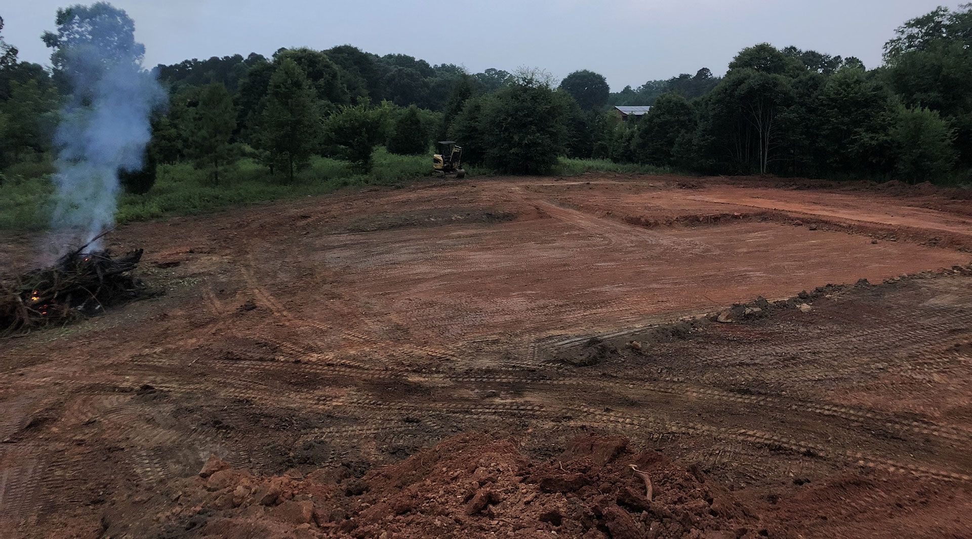 Cleared reddish dirt field with a small smoke plume on the left and trees lining the background