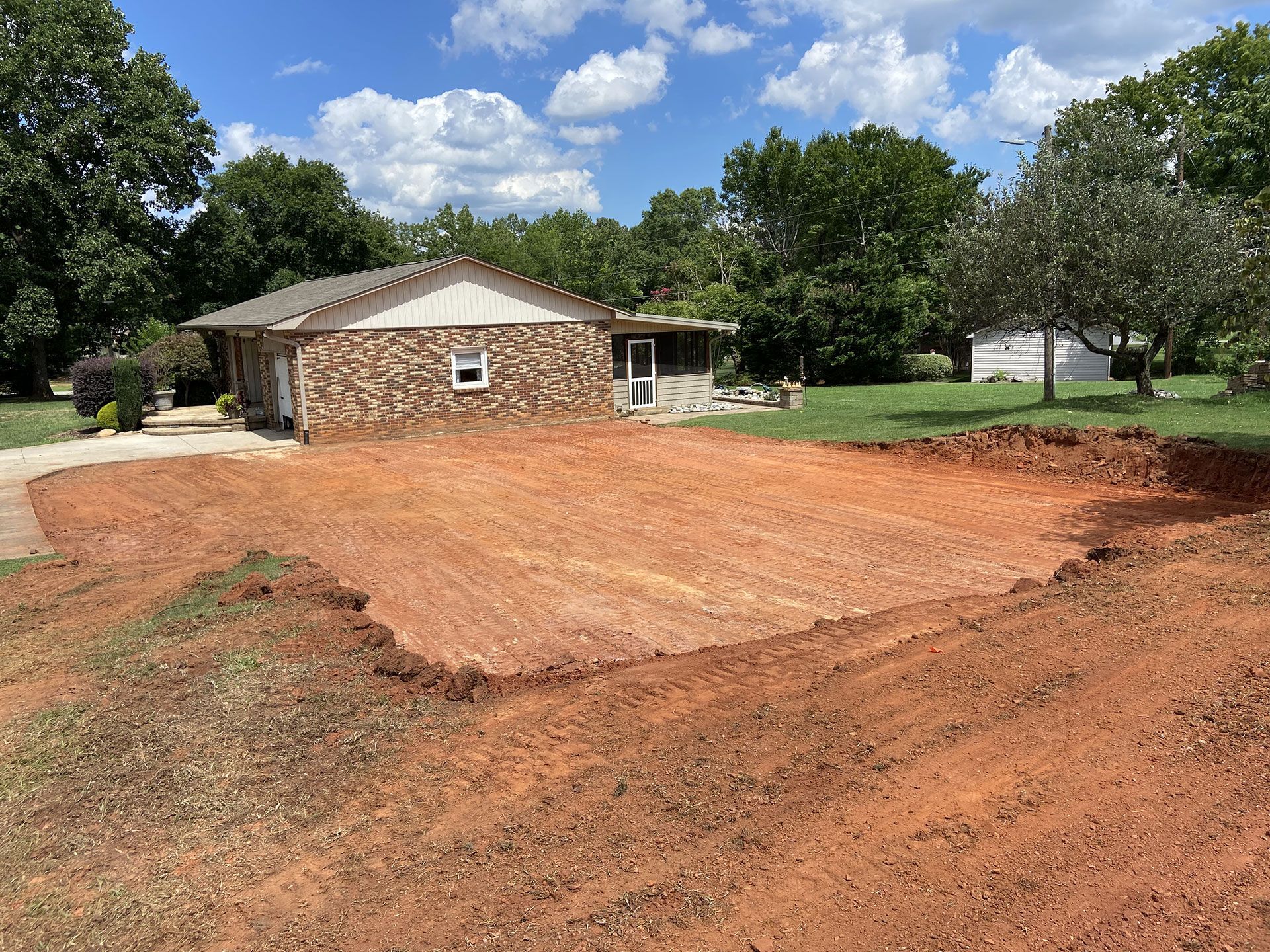 Freshly cleared red dirt lot beside a stone house under a blue sky with trees