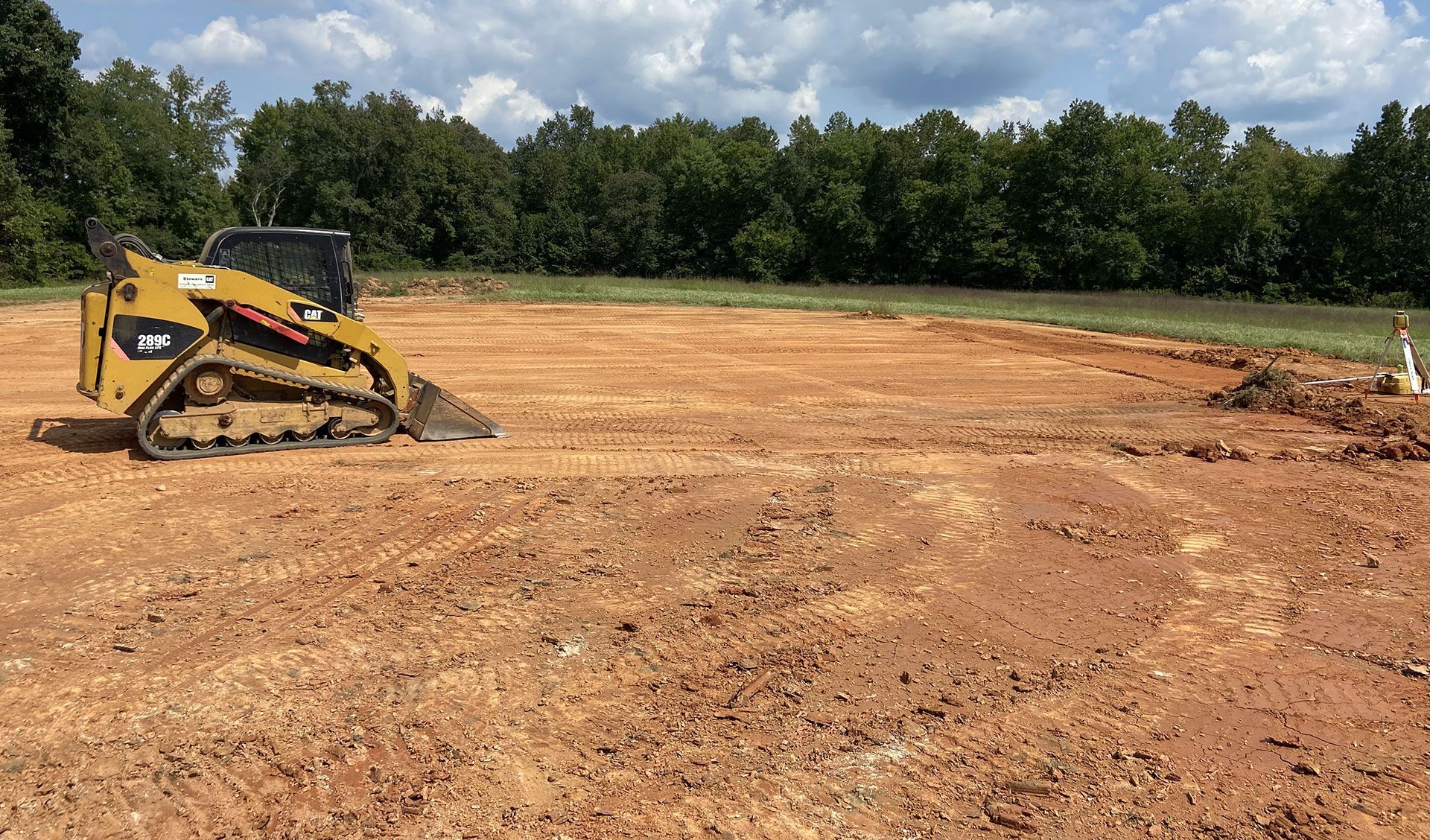 Yellow skid steer on a cleared dirt lot with trees and cloudy sky in the background