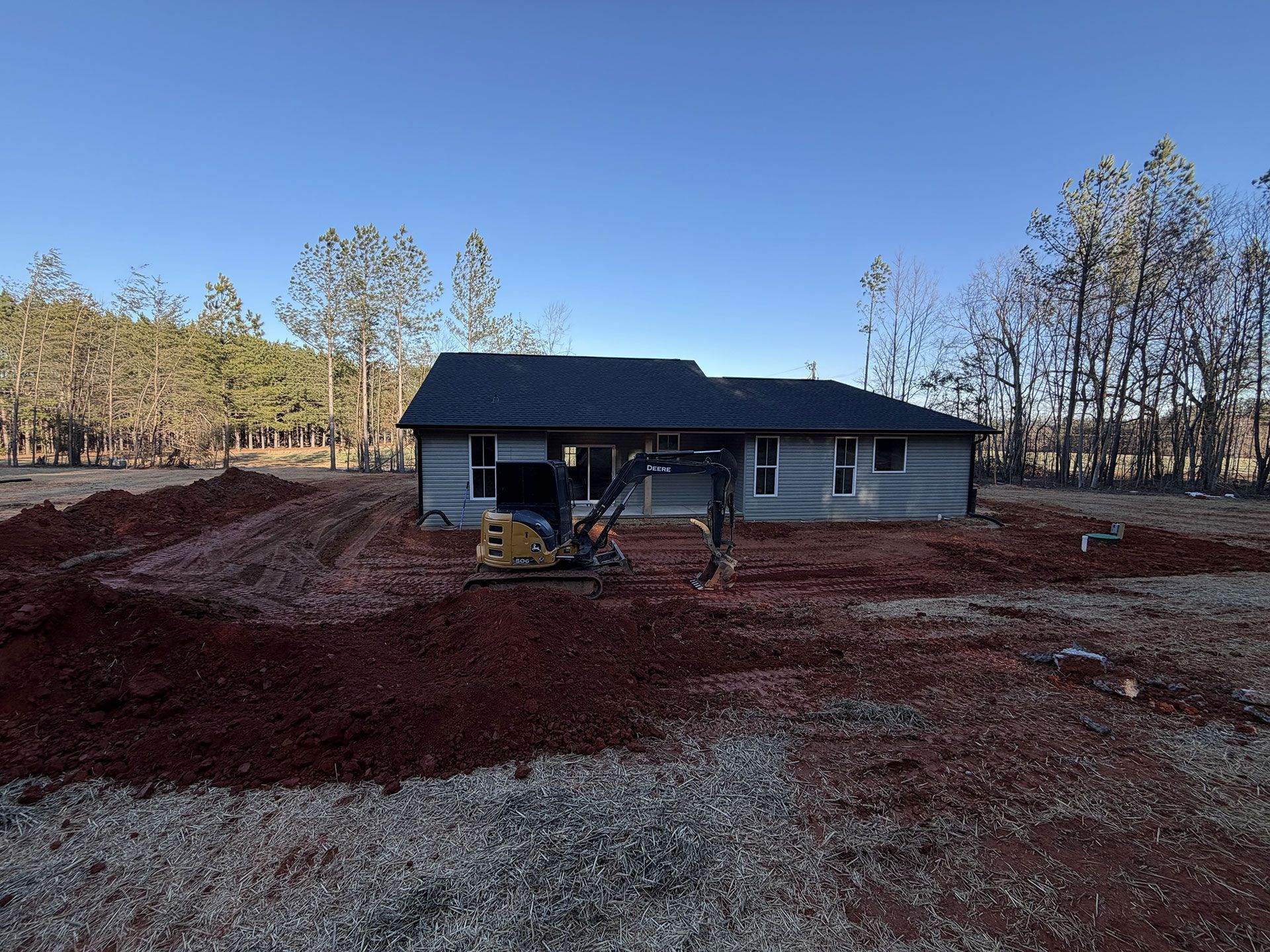 Single-story gray house under construction on a red dirt lot with a wooded background.