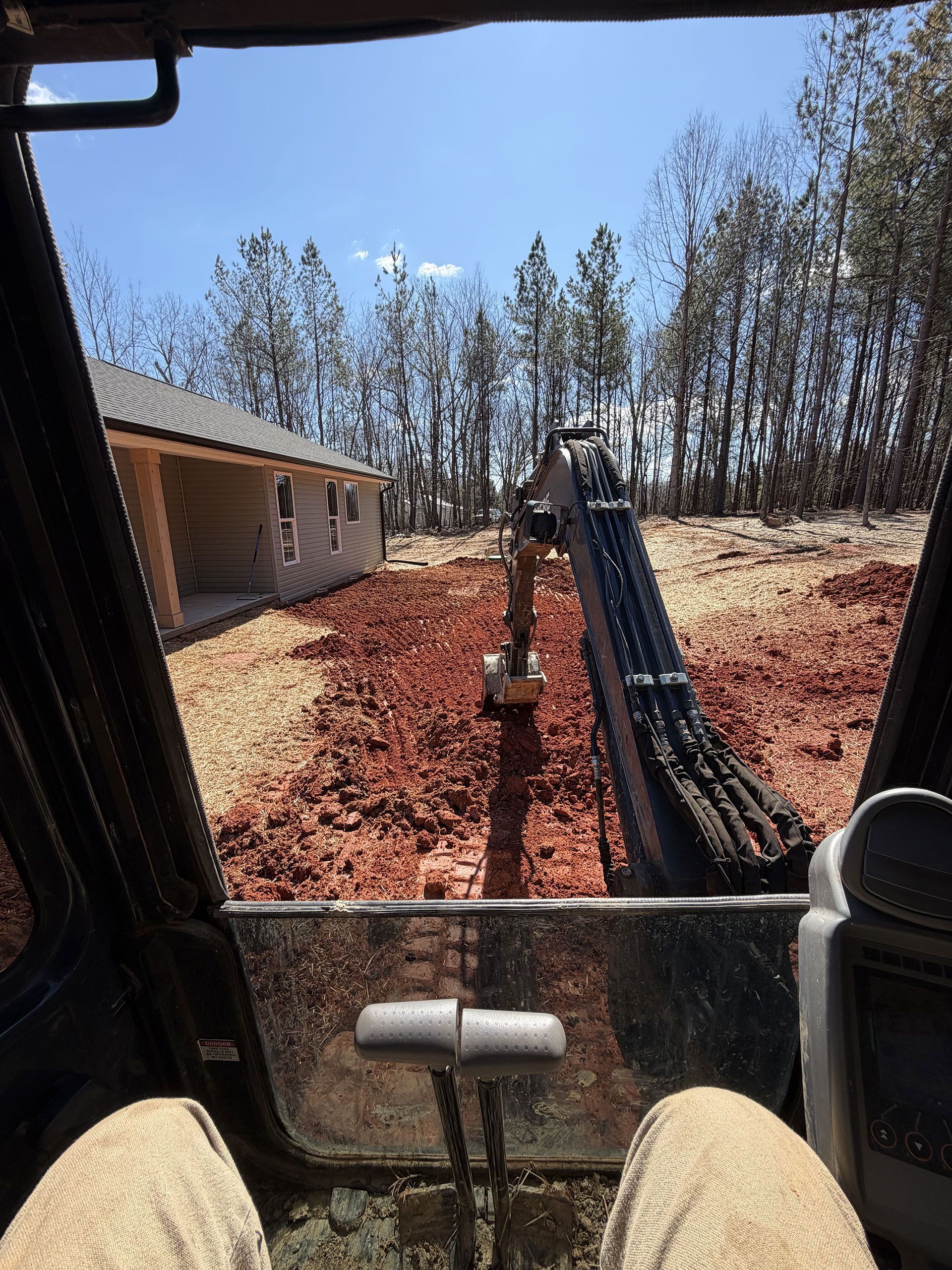 View from excavator cab of a dirt trench beside a house, with black pipes laid along the trench.