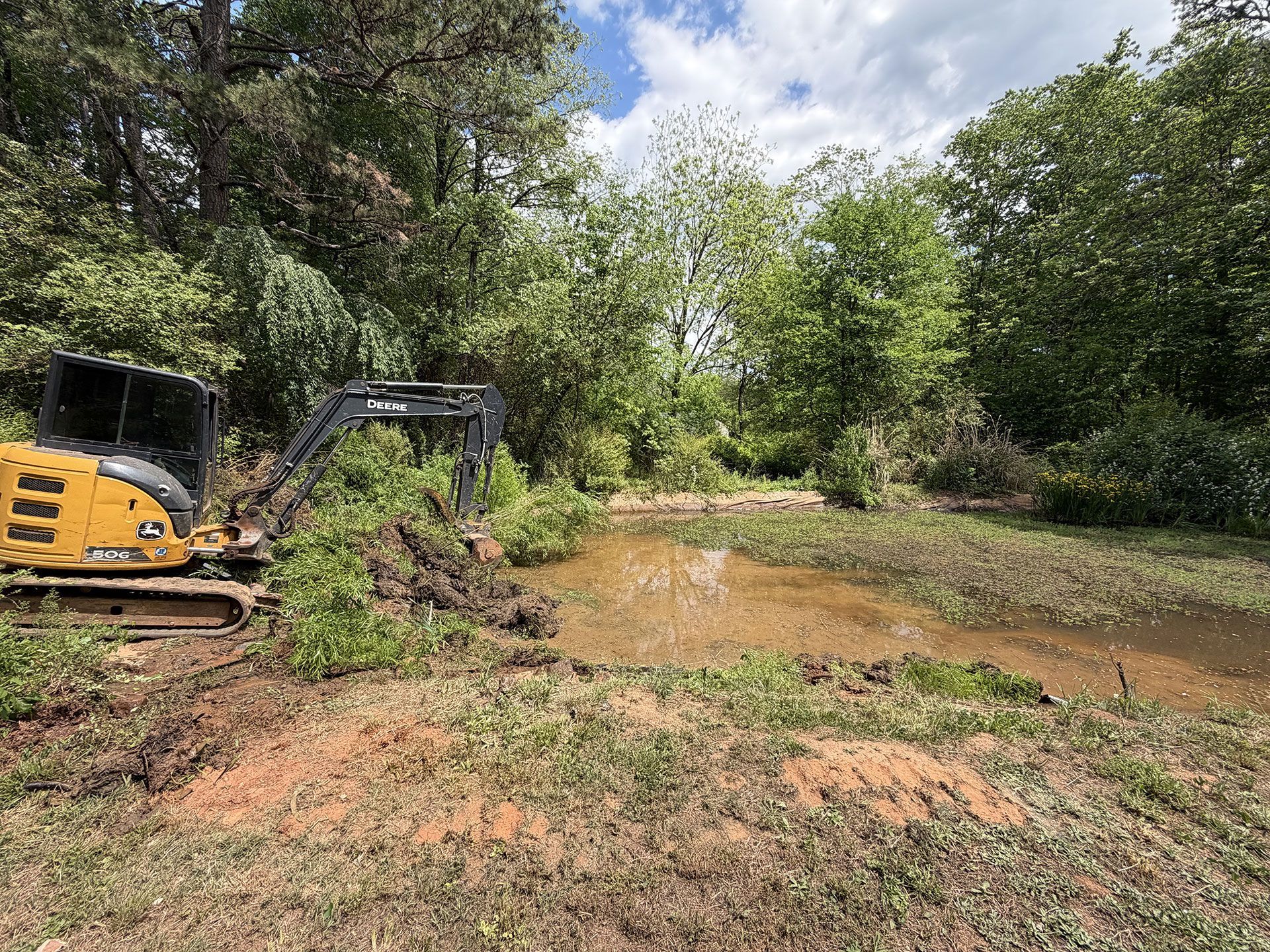 Yellow excavator clearing muddy ground beside a wooded pond under a cloudy sky