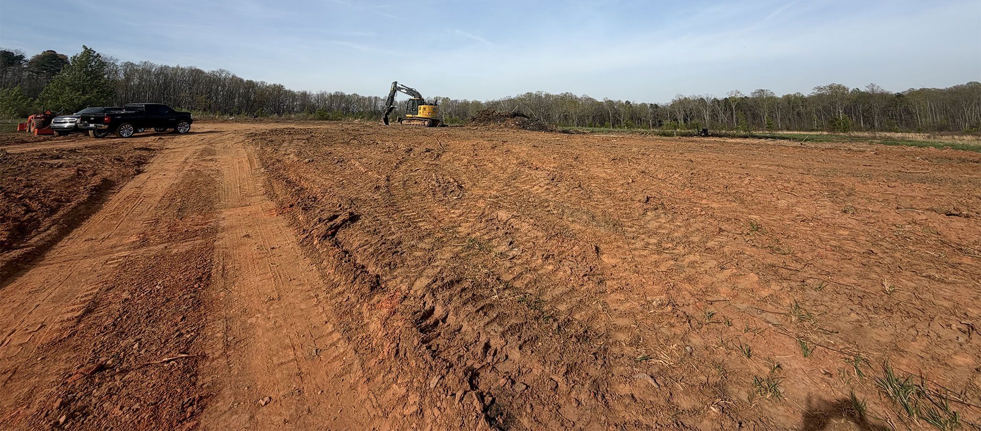 Red dirt field with a graded track, a tractor in the distance, and trees along the horizon.
