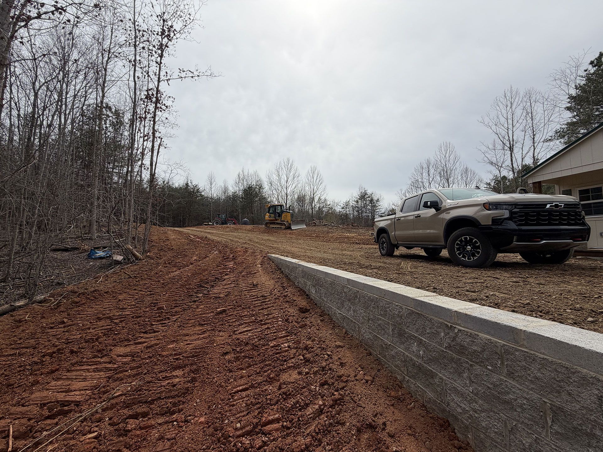 Concrete retaining wall beside dirt lot with parked pickup truck and wooded hillside under cloudy sky