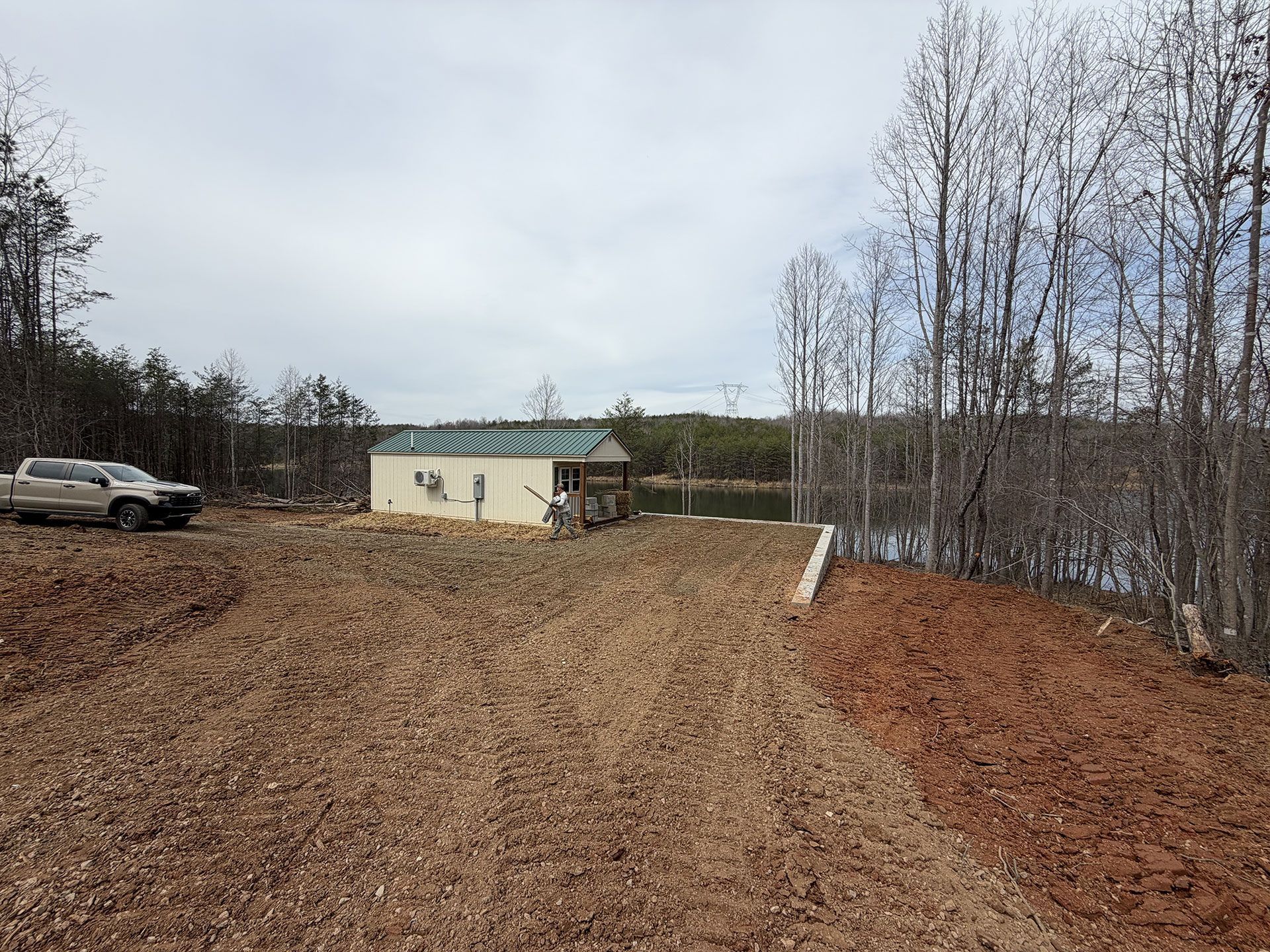 Dirt driveway leading to a small beige building, with a parked truck and leafless trees under a gray sky.