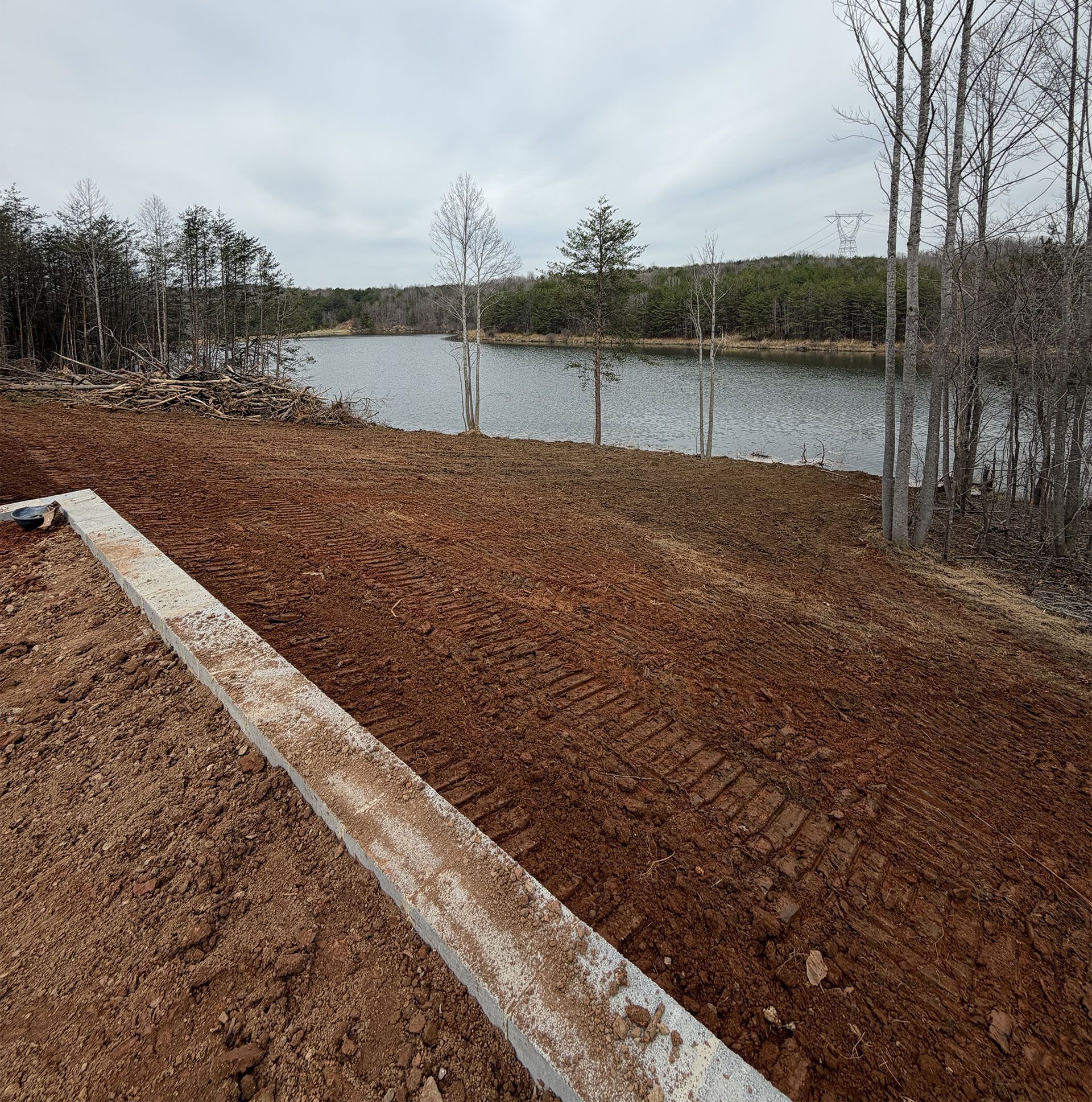 Dirt overlook by a lake under overcast sky, with bare trees and a wooden beam in the foreground