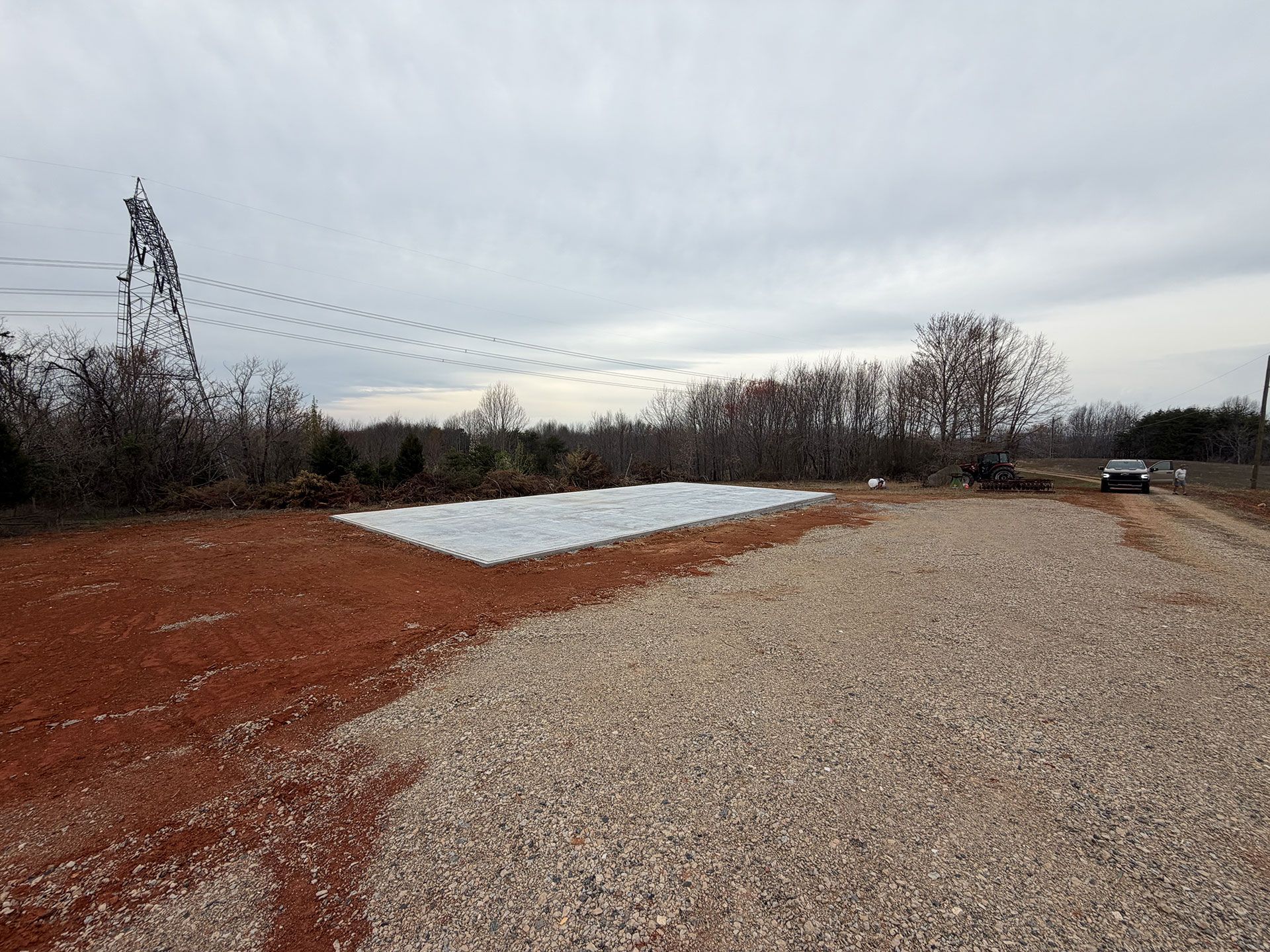 Wide gravel lot under cloudy sky with a white tarp and reddish dirt edges, bordered by bare trees.