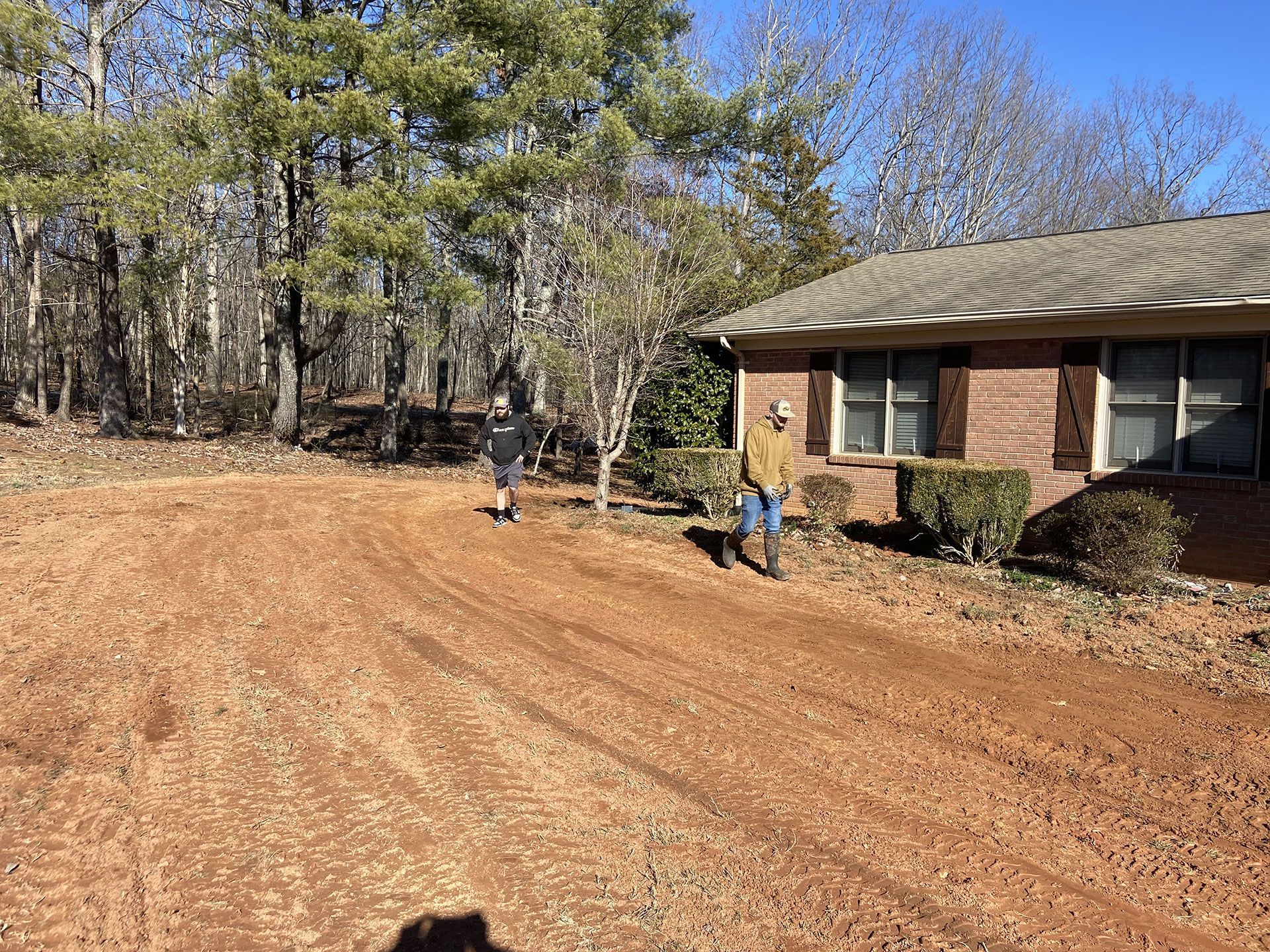 Dirt driveway beside a brick house, with two people near the trees in a wooded yard.