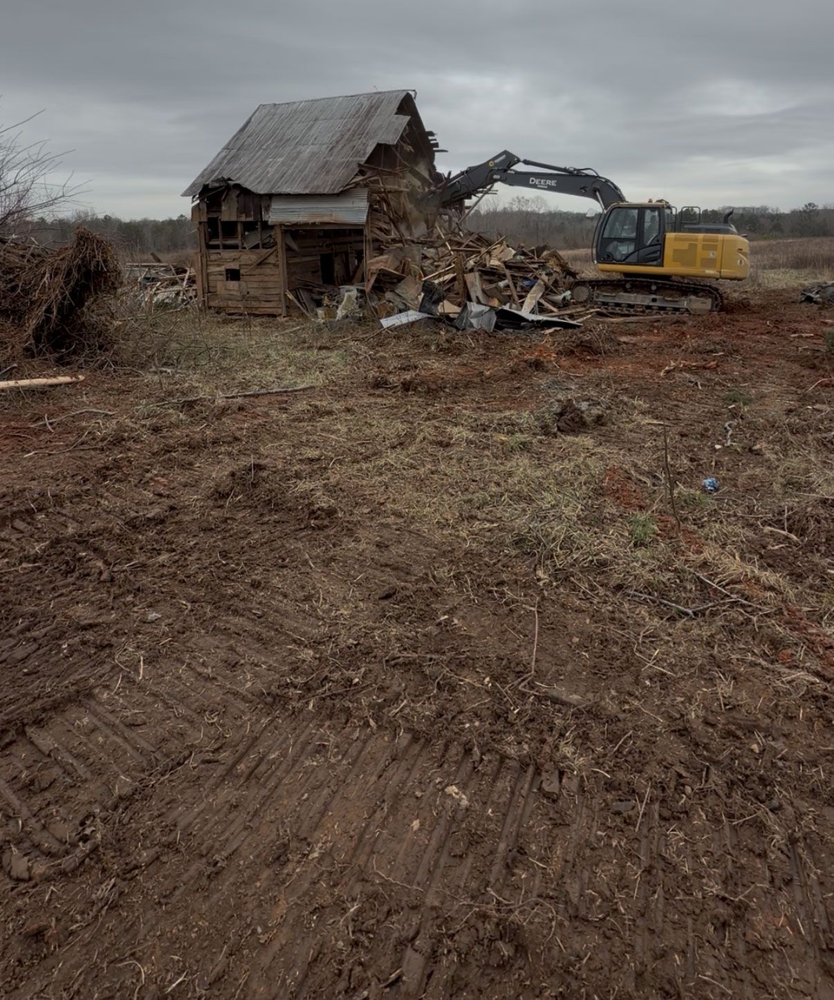 Excavator demolishing an old house in a muddy field under a gray sky