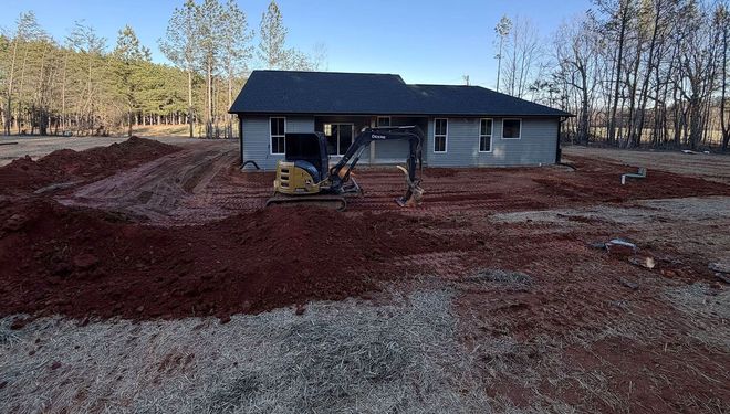 Single-story house with a dark roof, surrounded by red dirt in a wooded rural lot.
