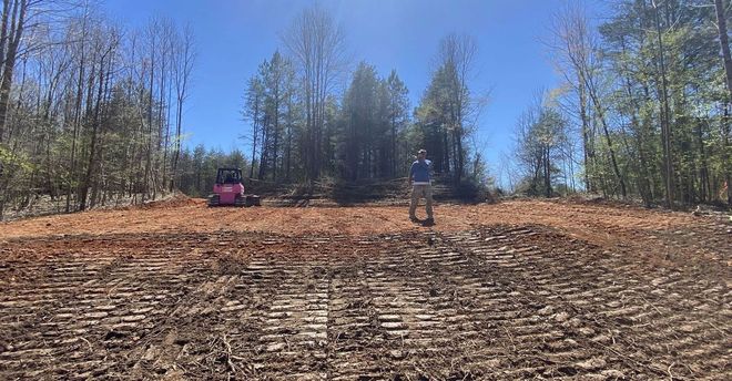 Cleared hillside with two workers and a red machine near a tree line under a blue sky