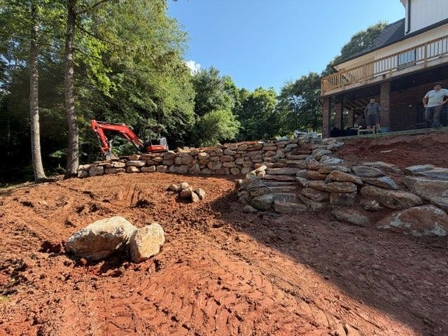 Construction site with red excavator, stone retaining wall, and dirt grading beside a house on a wooded slope