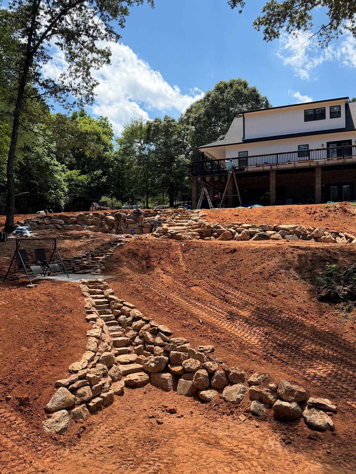 Reddish dirt yard with a stone path leading to a house under construction, surrounded by trees.